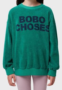 Niño vestido con un sudadera de forro polar verde con el texto "BOBO CHOSES" en azul oscuro y pantalones lilas claros.