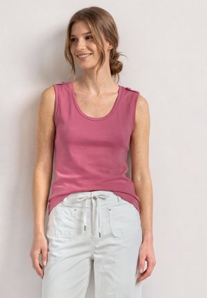 Femme aux cheveux châtain clair en chignon bas, portant un haut rose sans manches et un pantalon blanc à cordon, souriante et regardant sur le côté.