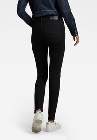 Schwarze skinny Jeans mit glatter Textur, hoher Taille und auffälligen Gesäßtaschen, kombiniert mit weißen Turnschuhen und einer Jeansjacke.