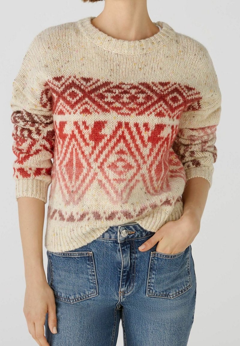 Femme portant un pull en maille crème et rouge à motifs, associé à un jean bleu taille haute avec des poches plaquées à l'avant.