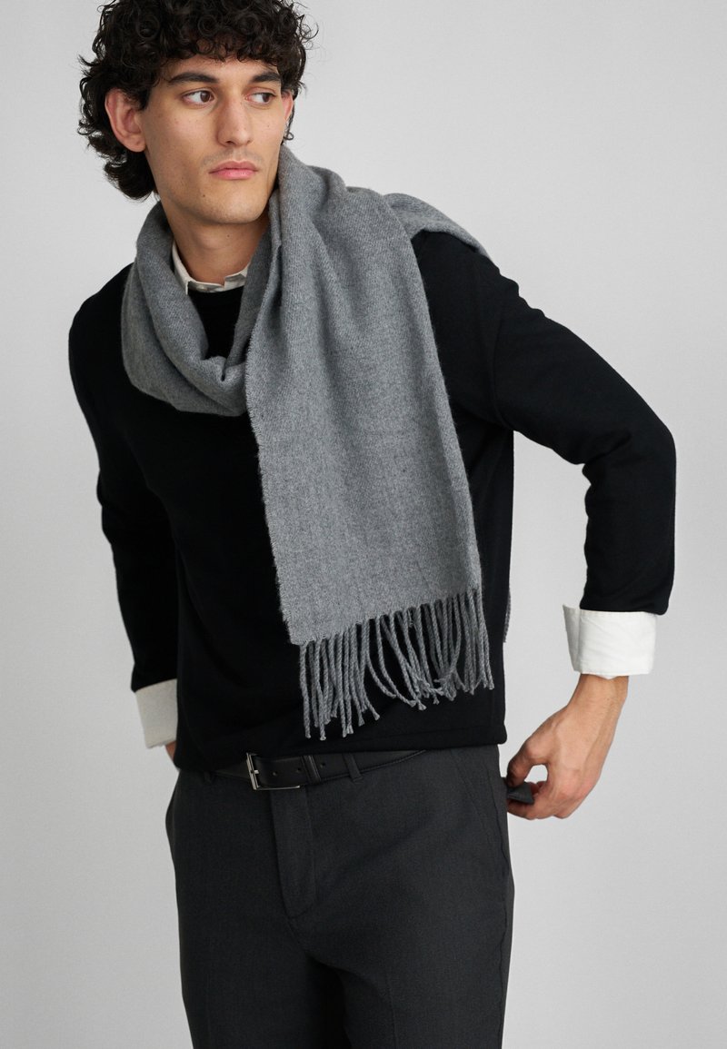 Écharpe grise avec des franges, faite d'un matériau doux, drapée autour du cou, associée à un pull noir et un pantalon gris.