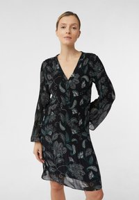 Eine Frau in einem schwarzen Kleid mit langen Ärmeln und blau-weißen Blumenmustern, die mit einer Hand auf der Hüfte vor einem schlichten Hintergrund steht.