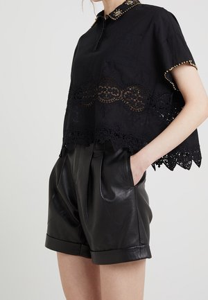 Kobieta w czarnej bluzce z krótkim rękawem wykończonej koronką, z kołnierzykiem ozdobionym złotymi ćwiekami oraz w czarnych, plisowanych skórzanych szortach z wysokim stanem i kieszeniami.
