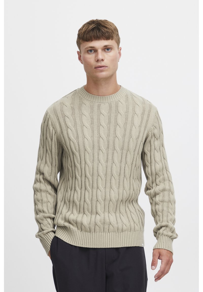 Pull en maille beige avec col rond, poignets et ourlet côtelés. Présente des motifs en câble texturés sur tout le corps.