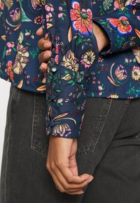 Blouse florale navy avec des motifs roses et verts vibrants, des poignets à boutons et un tissu doux et texturé. Le poignet présente un détail avec un onglet.