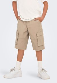 Shorts cargo beiges avec deux poches latérales, en tissu de mélange de coton. Portés avec un t-shirt blanc et des baskets blanches avec des chaussettes mi-mollet.