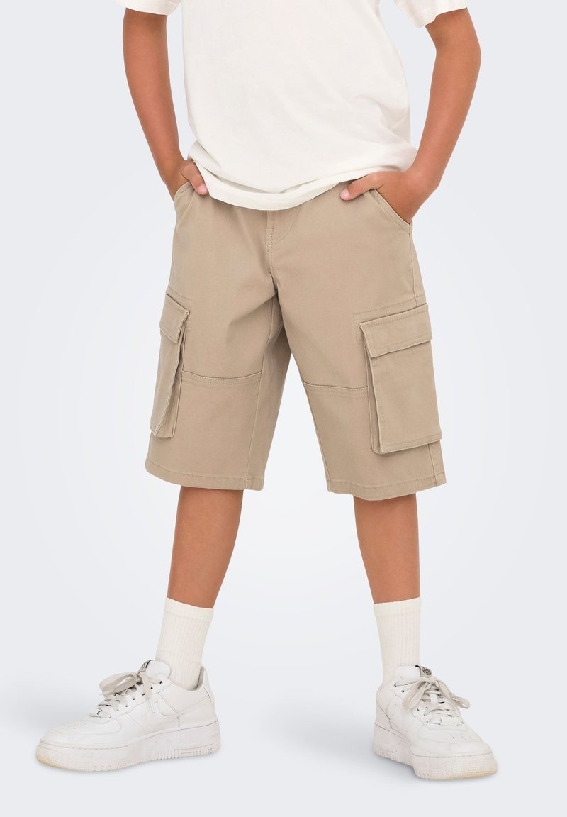 Shorts cargo beiges avec deux poches latérales, en tissu de mélange de coton. Portés avec un t-shirt blanc et des baskets blanches avec des chaussettes mi-mollet.
