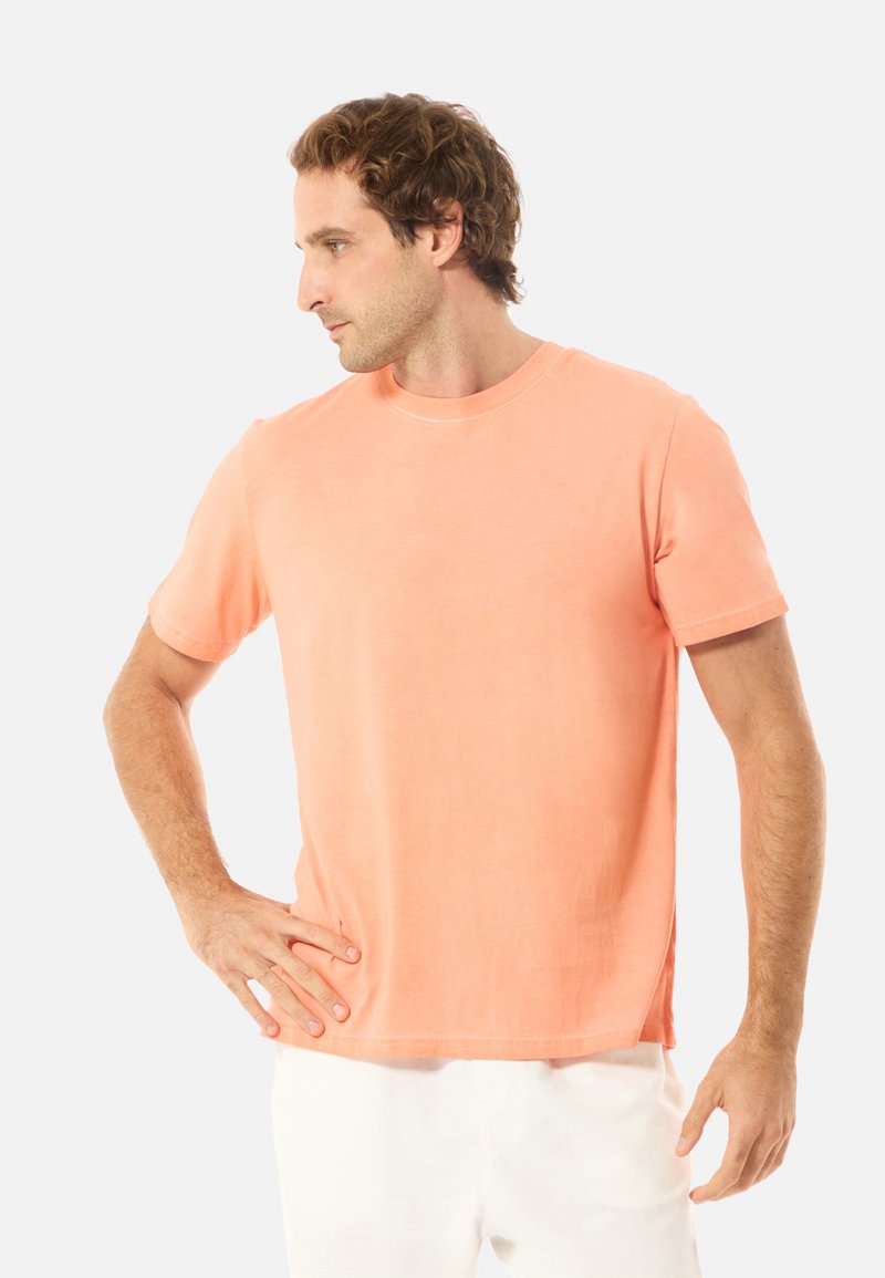 Homme portant un t-shirt corail à manches courtes et un pantalon blanc, debout avec une main sur la hanche, regardant vers la gauche sur un fond clair uni.