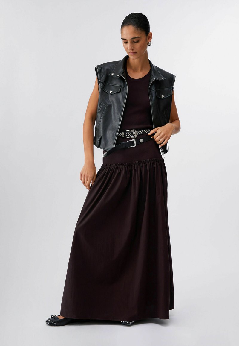 Woman with dark hair in a bun wearing a black sleeveless leather vest, a black sleeveless top, long black skirt, and studded belt and shoes.