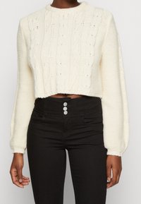 Vit croppad tröja med kabelstickad textur och långa ärmar, tillsammans med högmidjade svarta jeans med blanka silverknappar.