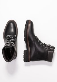 Bottes en cuir noir au design robuste, lacets gris et accessoires métalliques. Semelle épaisse à crampons pour une bonne adhérence, avec une hauteur classique à la cheville.