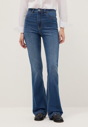 Femme portant un jean évasé bleu taille haute, une chemise blanche boutonnée et des mocassins noirs, debout devant un fond uni.