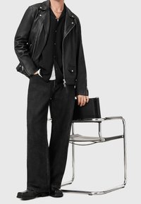 Giacca da motociclista nera in pelle, camicia nera, jeans neri a gamba larga e scarpe nere. La persona tiene una borsa nera, contro uno sfondo minimalista.