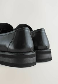 Schwarze Lederschuhe mit glänzender Oberfläche, runden Absätzen und einer dicken, schwarzen Gummisohle mit strukturiertem Streifendetail.