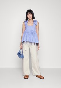 Mulher a usar uma blusa sem mangas azul e branca às riscas com laços, calças largas em tom creme, sandálias castanhas e a segurar uma pequena mala azul.