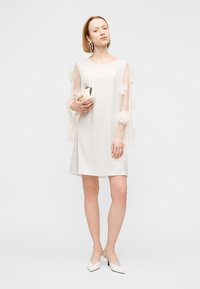 Άσπρο μίνι φόρεμα με διαφανείς, βολάν μανίκια, λεία υφή, άνετη εφαρμογή, σε συνδυασμό με μπεζ τσάντα χειρός και άσπρα παπούτσια με μυτερή μύτη.