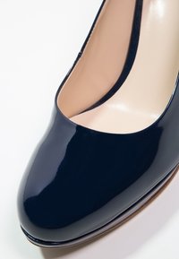 Escarpin pour femme bleu marine brillant à bout arrondi avec doublure intérieure beige sur fond blanc.
