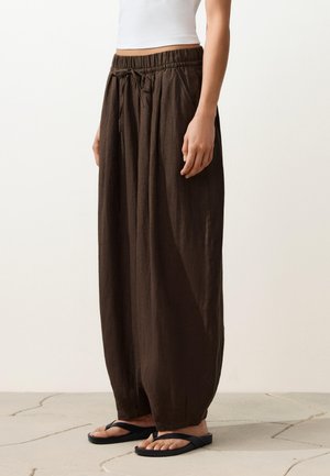 Vrouw draagt losse donkerbruine wijde broek met trekkoord in de taille, wit crop top en zwarte slippers, staand op betegelde vloer.