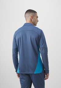 Majica z dolgimi rokavi športnega sloga, ki se ponaša z modro in turkizno barvno shemo, strukturirano tkanino ter ovratnikom s subtilno vzorčno podrobnostjo.