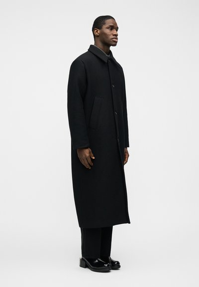 Άνδρας που στέκεται, φοράει μακρύ μαύρο παλτό με κουμπιά, μαύρο παντελόνι και μαύρα παπούτσια με τακούνι, μπροστά σε απλο φόντο φωτός.