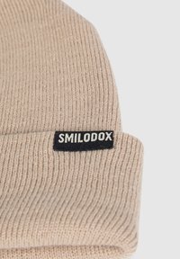 Beige knitted beanie with ribbed texture. Features a black fabric tag with the text "SMILODOX" in white.