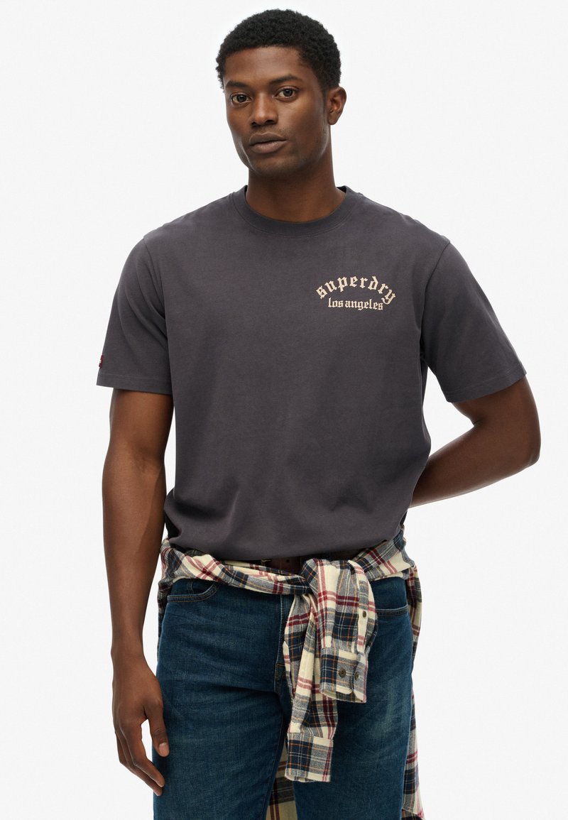 Sort bomulds T-shirt med korte Ærmer, med cremefarvet "Superdry Los Angeles" tekst. Mørkeblå jeans med en ternet skjorte bundet om livet.
