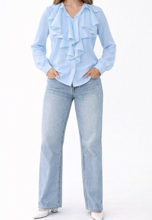 Kobieta w jasnoniebieskiej bluzce z falbankami, jasnoniebieskie proste jeansy, białe buty na niskim obcasie, ręce w kieszeniach, stoi na białym tle.