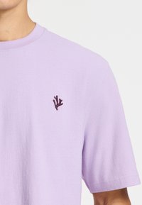 Lilas spalvos medvilninė marškinė su trumpomis rankovėmis. Yra vyno raudonos spalvos siuvinėti koraliniai raštai ant krūtinės. Švelni tekstūra, klasikinio apvalaus kaklo dizainas.