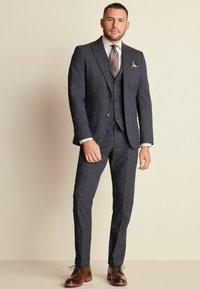 Traje de tres piezas azul oscuro con solapas de muesca, camisa gris claro y corbata estampada. Zapatos de vestir marrones completan el conjunto.
