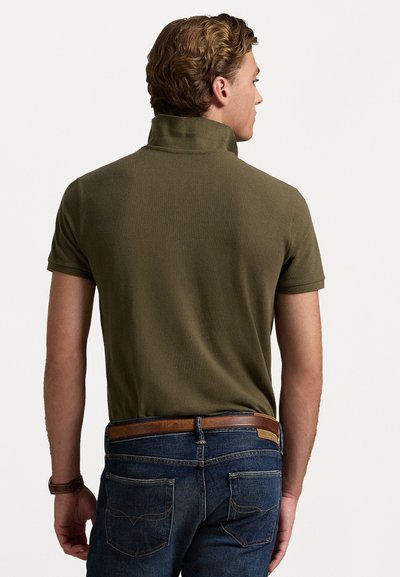Olive roheline polo särk, millel on krae, lühikesed varrukad ja tekstureeritud kangas. Kandnud tumedate siniste teksade ja pruuni nahkvööga.