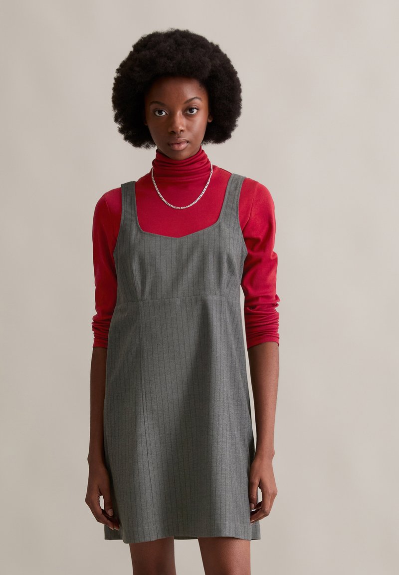 Grau gestreiftes Kleid mit quadratischem Ausschnitt, über einem enganliegenden roten Rollkragenpullover getragen. Texturiert mit entspannter Passform. Akzentuierte silberne Kettenhalskette.
