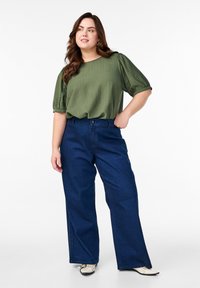 Blouse texturée vert olive avec des manches ballons, associée à un pantalon en denim large bleu. Le mannequin porte des chaussures blanches avec des accents noirs.