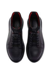 Un paio di scarpe nere in pelle con lacci, con inner lining rosso e lati testurizzati, viste dall'alto, su uno sfondo bianco.