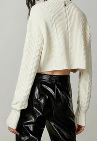 Valkoinen lyhyt neulepusero ribbineuleella ja palmikkokuvioinnilla; siinä on pitkät hihat, leveät hihansuut ja hopeinen vetoketju takana. Yhdistetty mustiin nahkahousuihin.