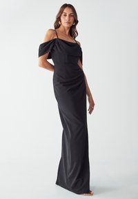 Abito da sera nero con maniche off-the-shoulder, corpetto aderente e dettagli drappeggiati lungo la vita. Finitura in satin liscio. Lunghezza lunga.