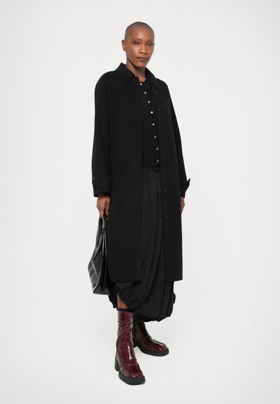 Abrigo largo negro, cierre de botones, combinado con una blusa negra y una falda texturizada. Botas de charol burdeos. Bolsa negra con un acabado liso.