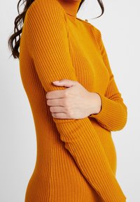 Rybrowana sukienka z golfem w intensywnym pomarańczowym kolorze, o dopasowanej sylwetce i długich rękawach z fakturowanym wykończeniem oraz subtelnym detalem na ramionach.