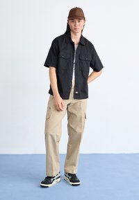 Sort kortærmet skjorte med to brystlommer, khaki cargo-bukser med sidelommer og sorte sneakers med hvide detaljer. Brun kasket.