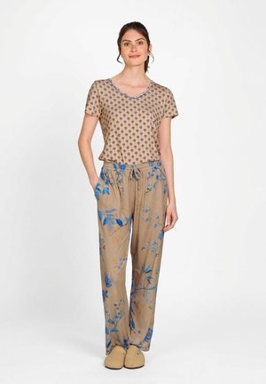 Vrouw staat met handen in haar zakken, draagt een beige top met korte mouwen en een klein bloemenpatroon, beige wijde broek met blauw bloemmotief en pantoffels.