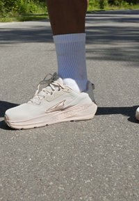 Light beige athletic shoes with textured material, lace-up design, and a thick sole. Worn with white ribbed socks on a concrete surface.