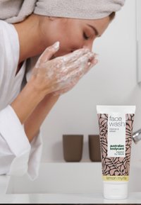 Vit tub med ansiktstvätt med rosa botanisk design, märkt "lemon myrtle." Person med handduk på huvudet löddrar ansiktet med tvål.