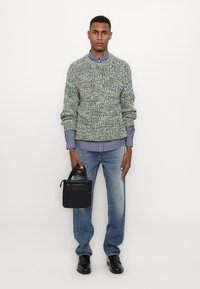 Maglione verde a maglia con un motivo texturizzato, indossato su una camicia a righe blu e bianche. Jeans azzurri chiari e una borsa nera con manici.