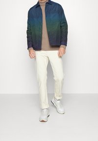 Man som bär en jacka med blå- till grönövergång över en beige polotröja, off-white byxor som är upploppade vid vristerna, och vita sneakers, stående på ett vitt golv.