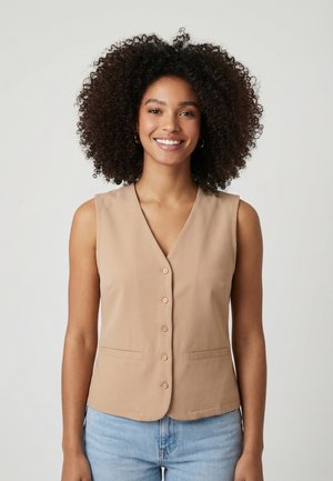 Donna sorridente con capelli ricci che indossa orecchini a cerchio, un gilet beige senza maniche con bottoni e jeans azzurri chiari su uno sfondo semplice.