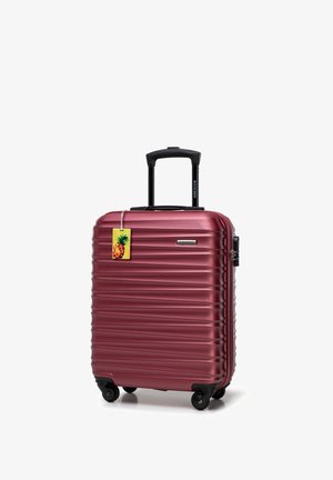 Valise rigide en métal bordeaux avec texture striée, poignée rétractable noire, quatre roues, et accentuée par une étiquette de bagages en forme d'ananas.