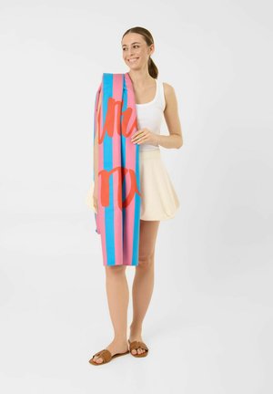 Femme souriante en débardeur blanc, jupe crème et sandales marron tenant une serviette rayée rose et bleue avec des lettres rouges.