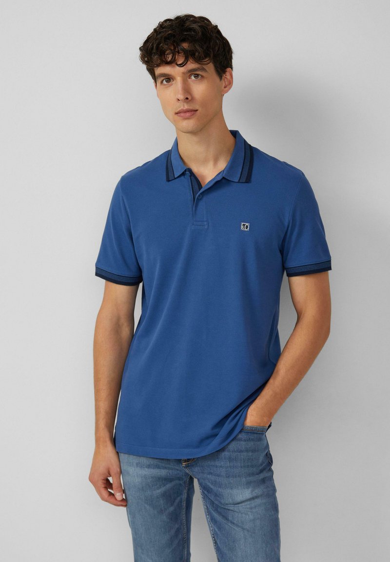 Niebieska koszulka polo wykonana z bawełny, z kołnierzykiem i granatowymi paskami na kołnierzu oraz rękawach, w zestawieniu z niebieskimi dżinsami.