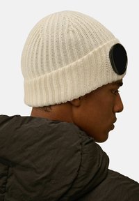 Beanie lavorato a maglia color crema con un motivo a coste testurizzato, caratterizzato da un accento circolare nero sulla parte frontale. Indossato sopra una giacca scura.