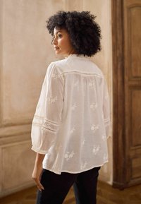 Hvid broderet bluse med puffede ærmer, dekoreret med blomsterprint og blondeaccenter langs manchetterne og bagpå skulderpartiet. Blødt, let stof.