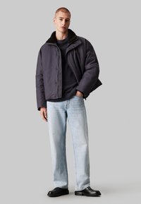 Puffjakke i mørk grå med en teksturert, svart saueskinnskrage. Brukt over en marineblå genser og kombinert med lyseblå vidde jeans og svarte sko.